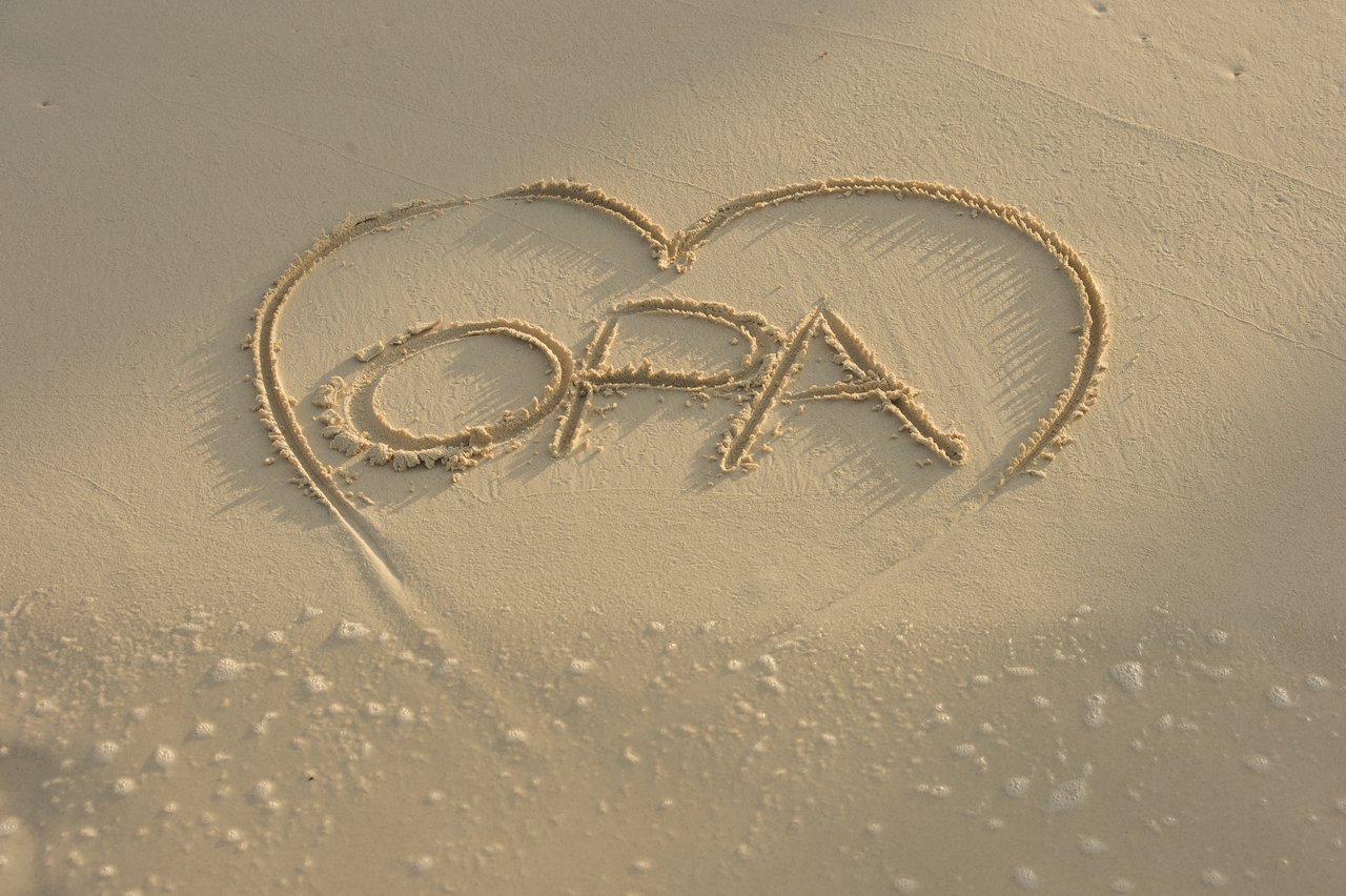 Heart shape drawn in sand with the word 'OPA' inside, near the water's edge.