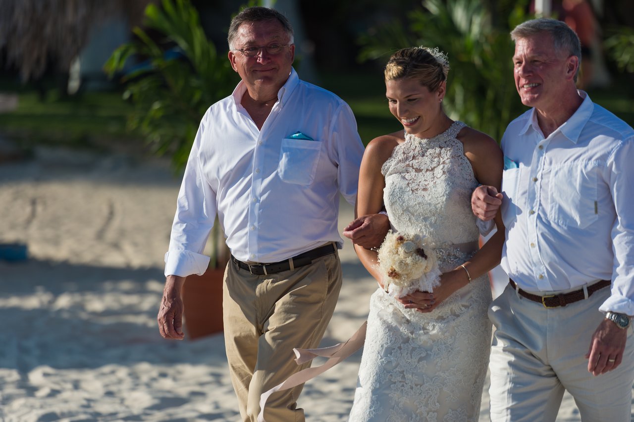A bride in a white dress walks on the beach, holding a bouquet and linking arms with two men.