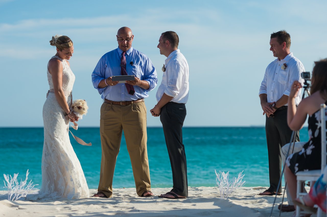 A bride and groom stand with an officiant on a beach during their wedding ceremony, with guests watching.