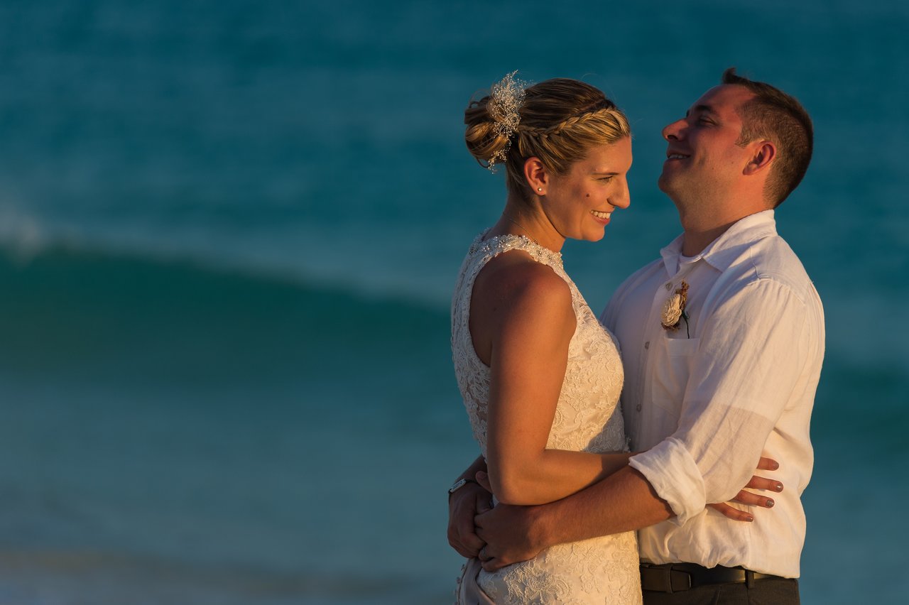 A bride and groom embrace and smile at each other on a beach during their wedding.