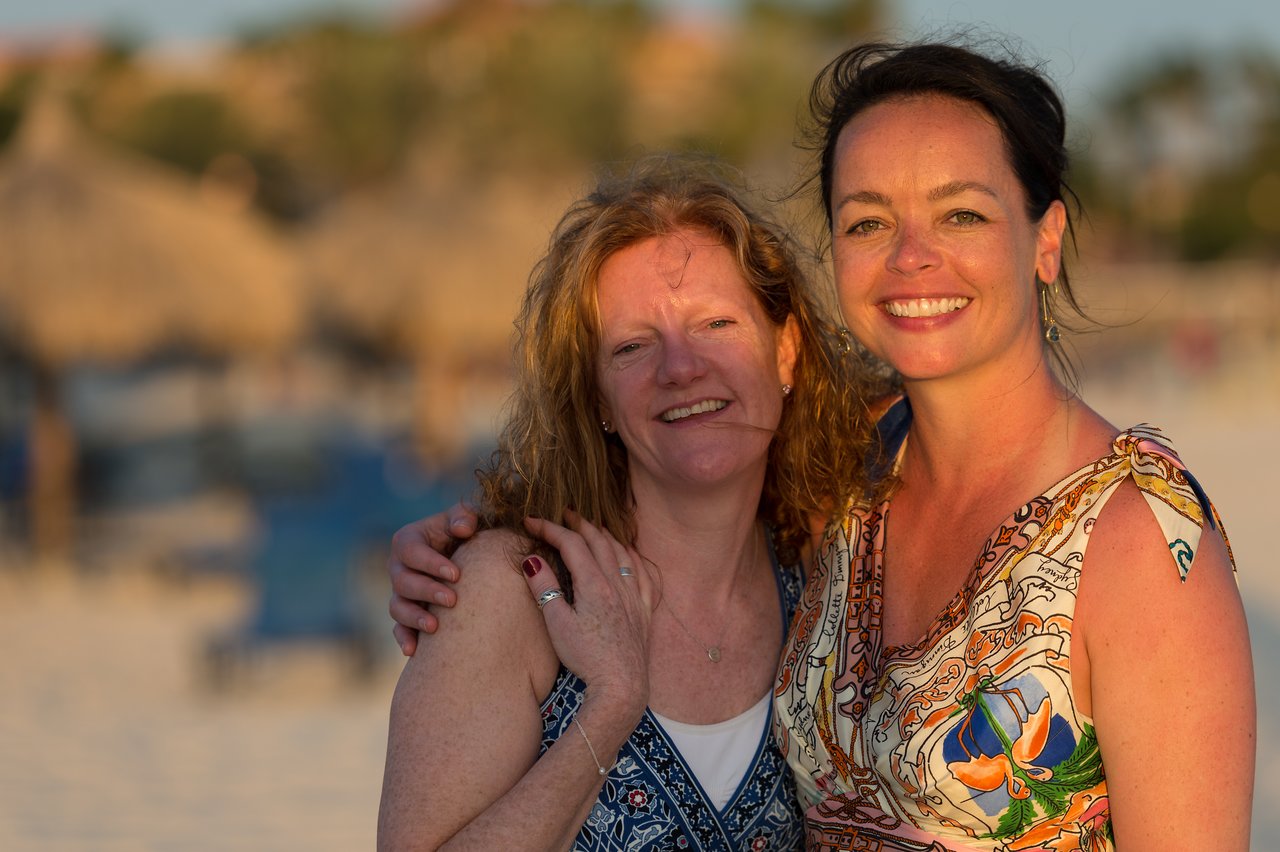 Two smiling women pose together on a beach, one with her arm around the other.