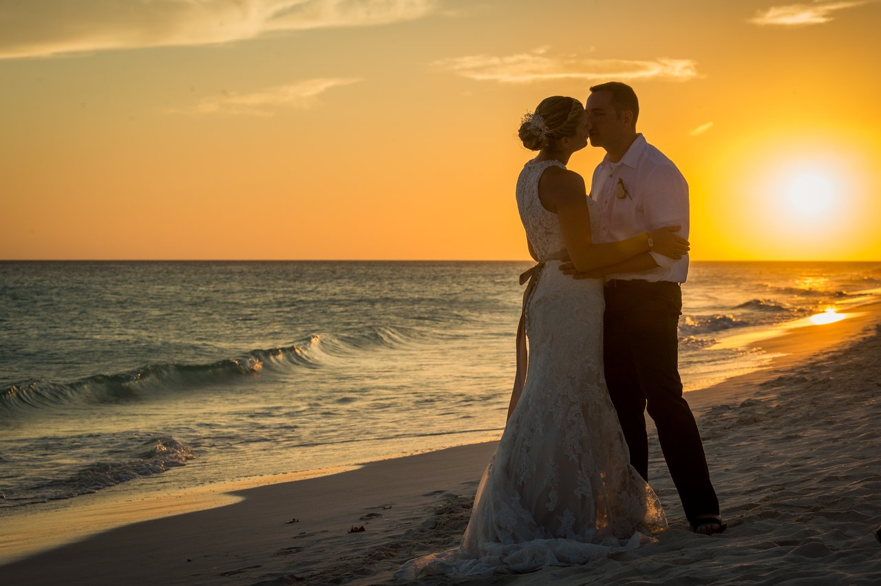 A bride and groom share a kiss on the beach at sunset.