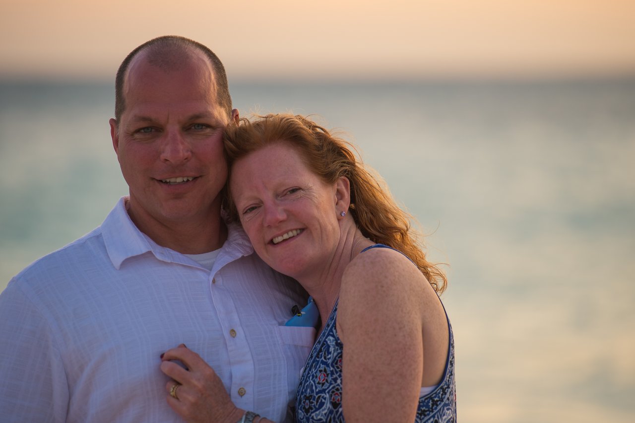 A smiling couple poses together near the ocean, with the woman leaning her head on the man's shoulder.