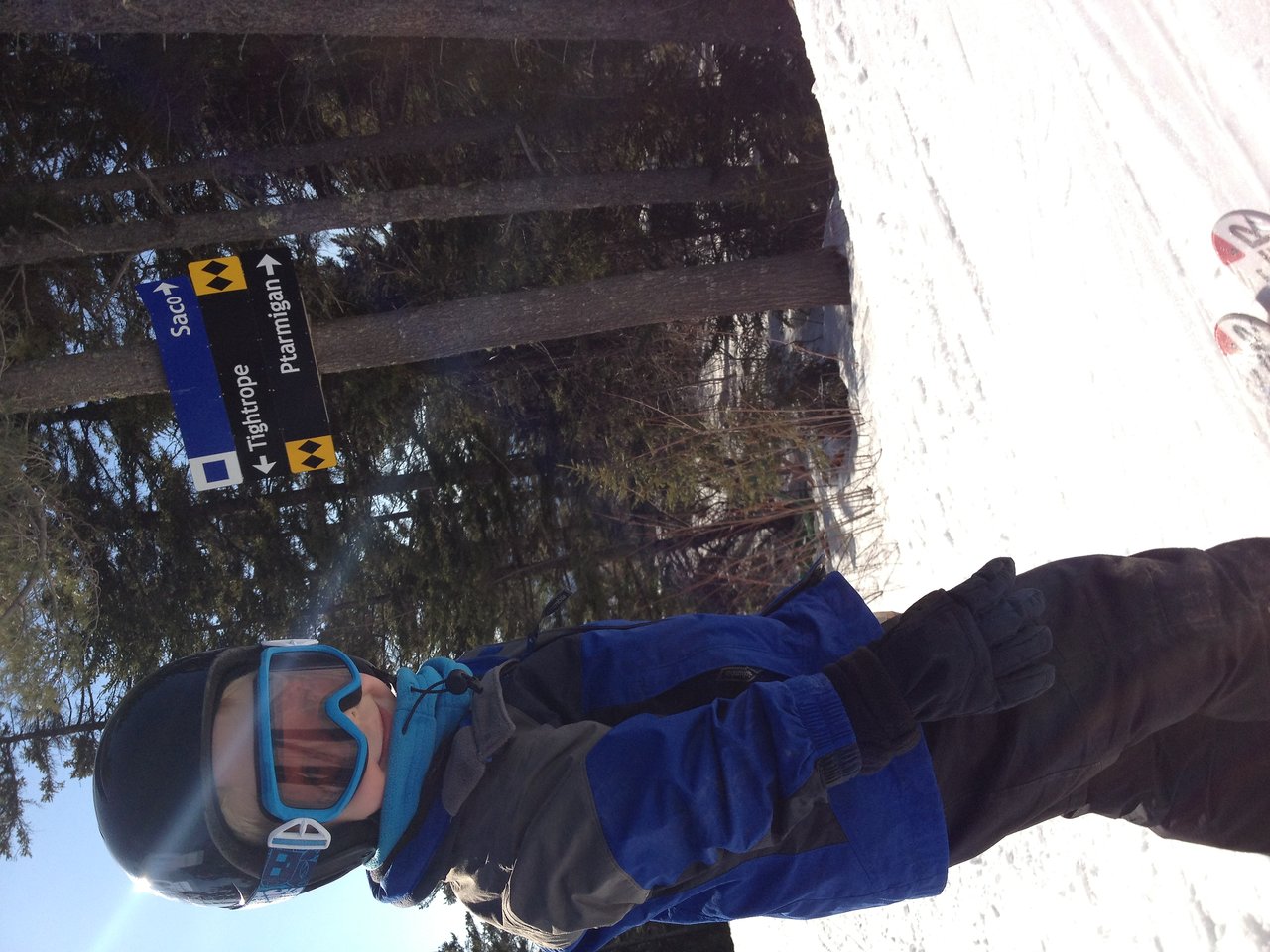 A child in a blue ski jacket and helmet stands on a snowy slope near a ski trail sign.