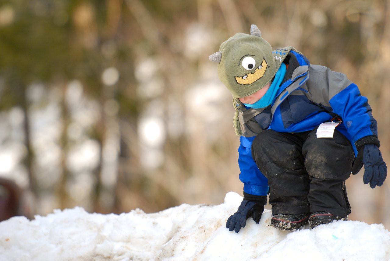 A child wearing a monster hat and winter clothes sits on a snowy surface, touching the snow with one hand.