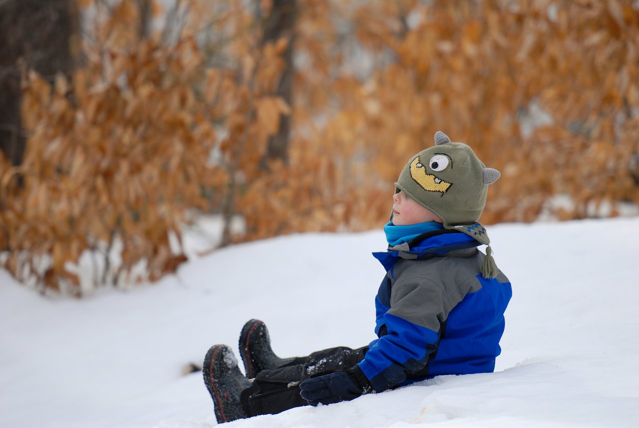 A child in a blue jacket and monster hat sits on the snow, looking to the side.