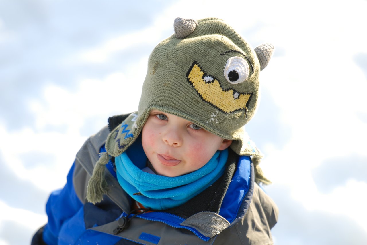 A child in a monster hat sticks out their tongue while wearing a winter coat and scarf in the snow.