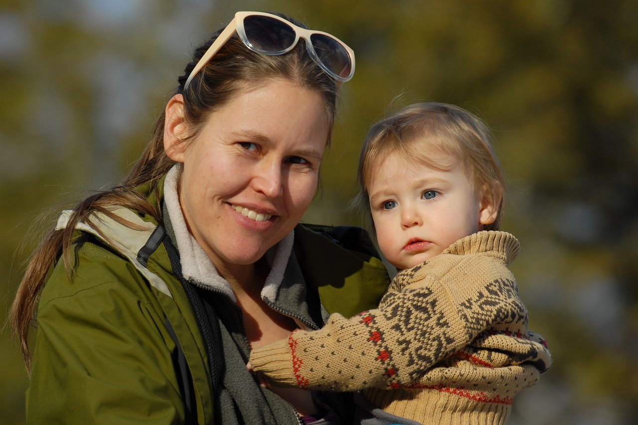 A woman in a green jacket holds a young child in a sweater while smiling at the camera.