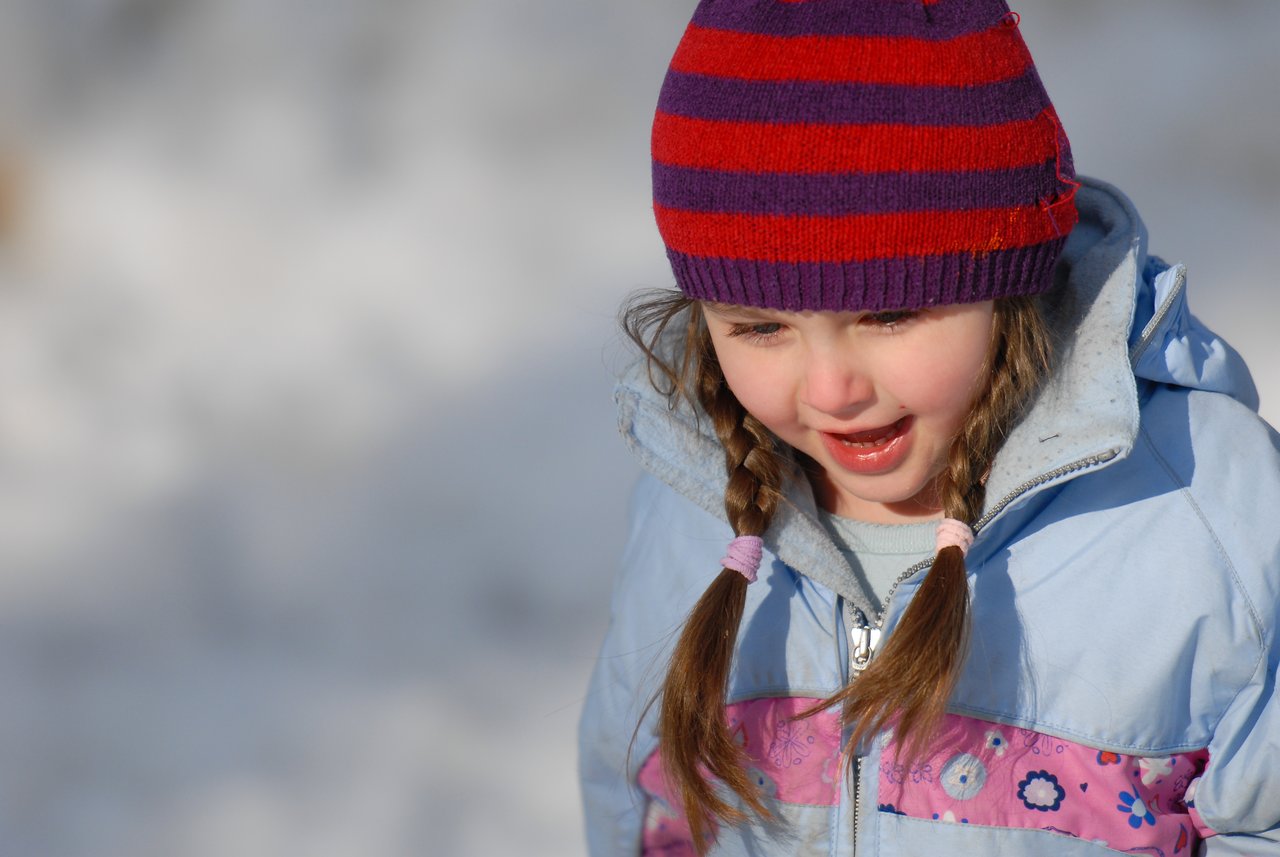A young child in a winter coat and striped hat smiles while playing outside in the snow.