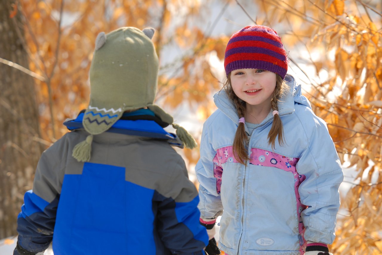 A young girl in a winter coat and hat smiles while standing outside with another child in the snow.