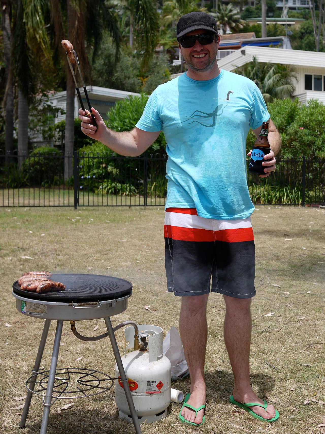 A man in sunglasses and a hat smiles while holding tongs with a sausage and a beer near a barbecue.