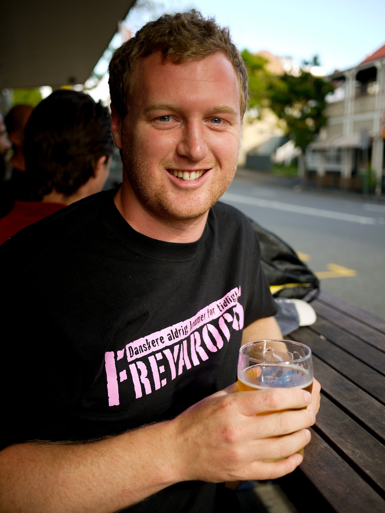 A man sits at an outdoor table, smiling and holding a glass of beer.