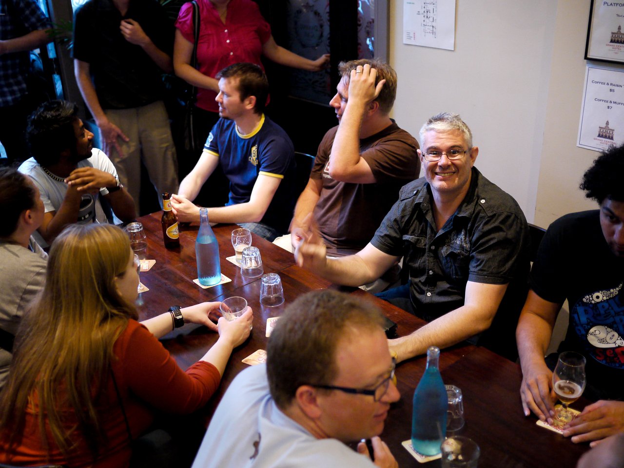 A group of people gathered around a table at a Brisbane Drupal meetup, talking and enjoying drinks.