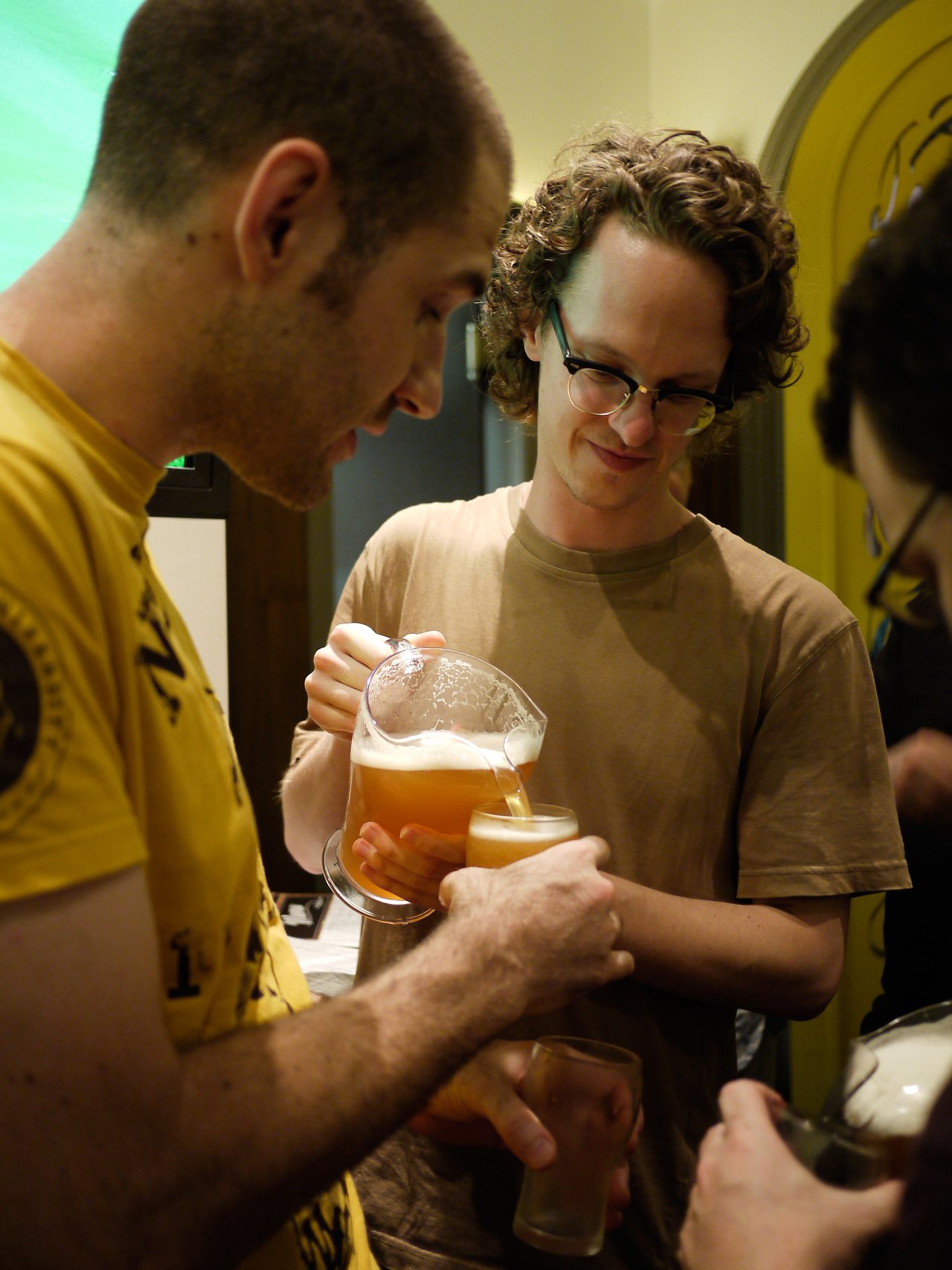 A man in glasses pours beer from a pitcher into a glass while chatting with others at a meetup.