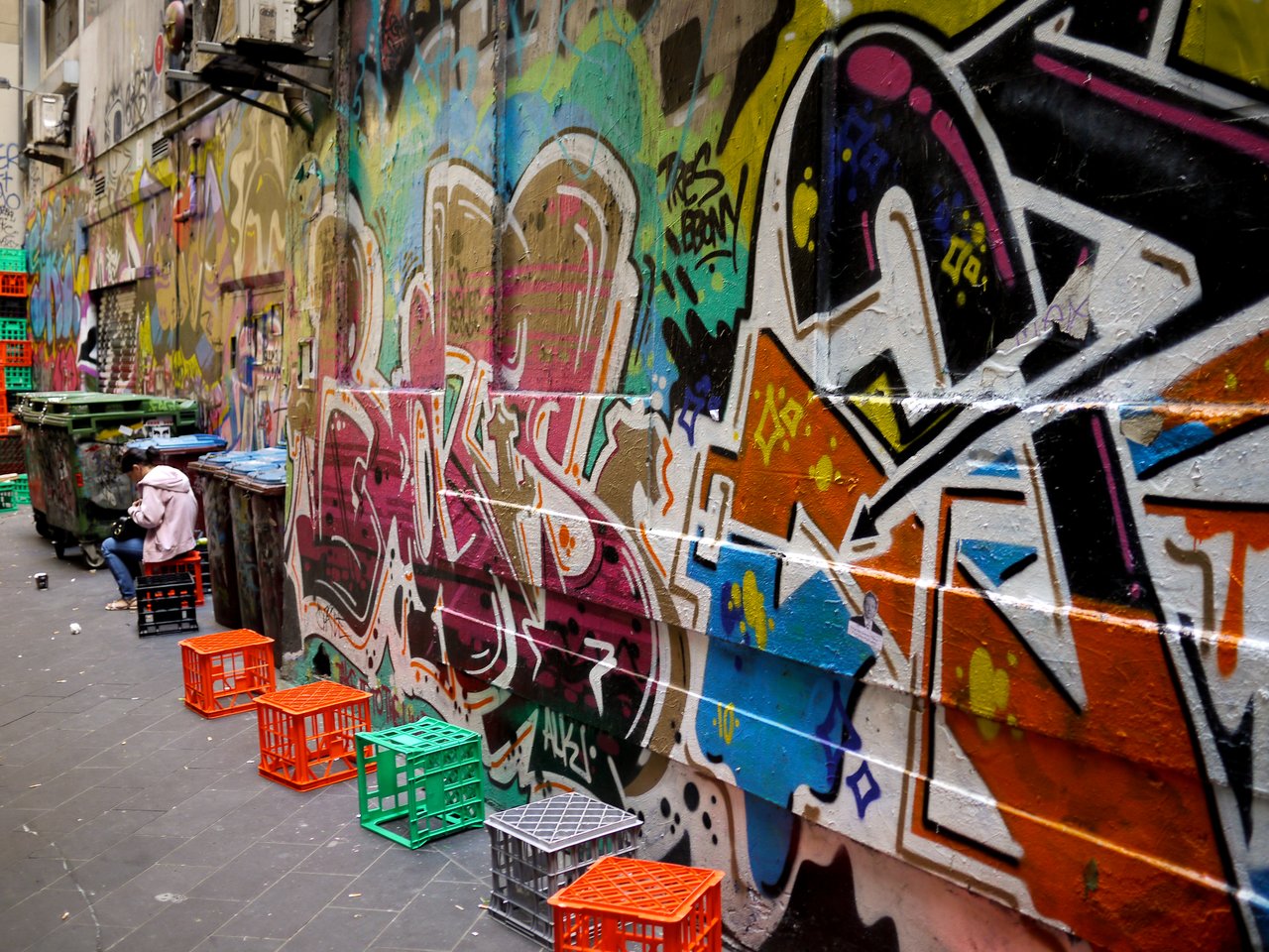 A graffiti-covered alley in Melbourne with colorful artwork on the walls and a person sitting on a crate.