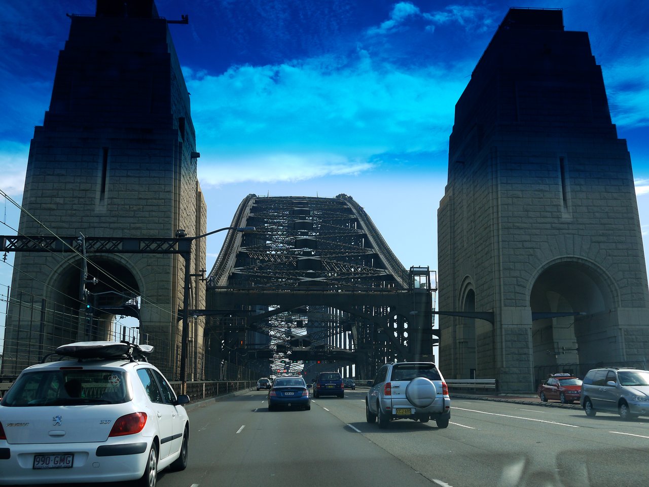 Cars driving on the Sydney Harbour Bridge, framed by two large stone pillars under a bright blue sky.