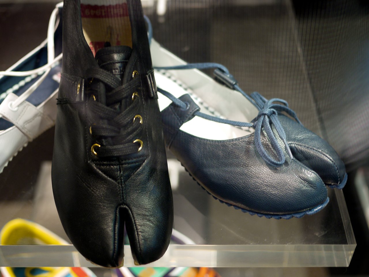 A pair of black and blue split-toe shoes resembling hooves displayed on a glass shelf.
