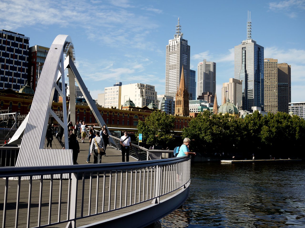 People walk across a modern bridge with Melbourne's skyline in the background, while one person leans on the railing.