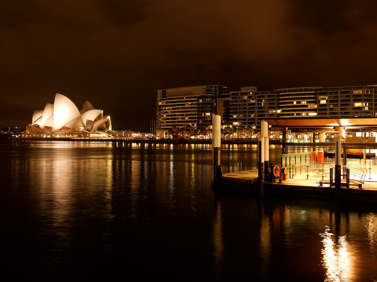 The Sydney Opera House is illuminated at night, reflecting on the water, with a dock and buildings nearby.