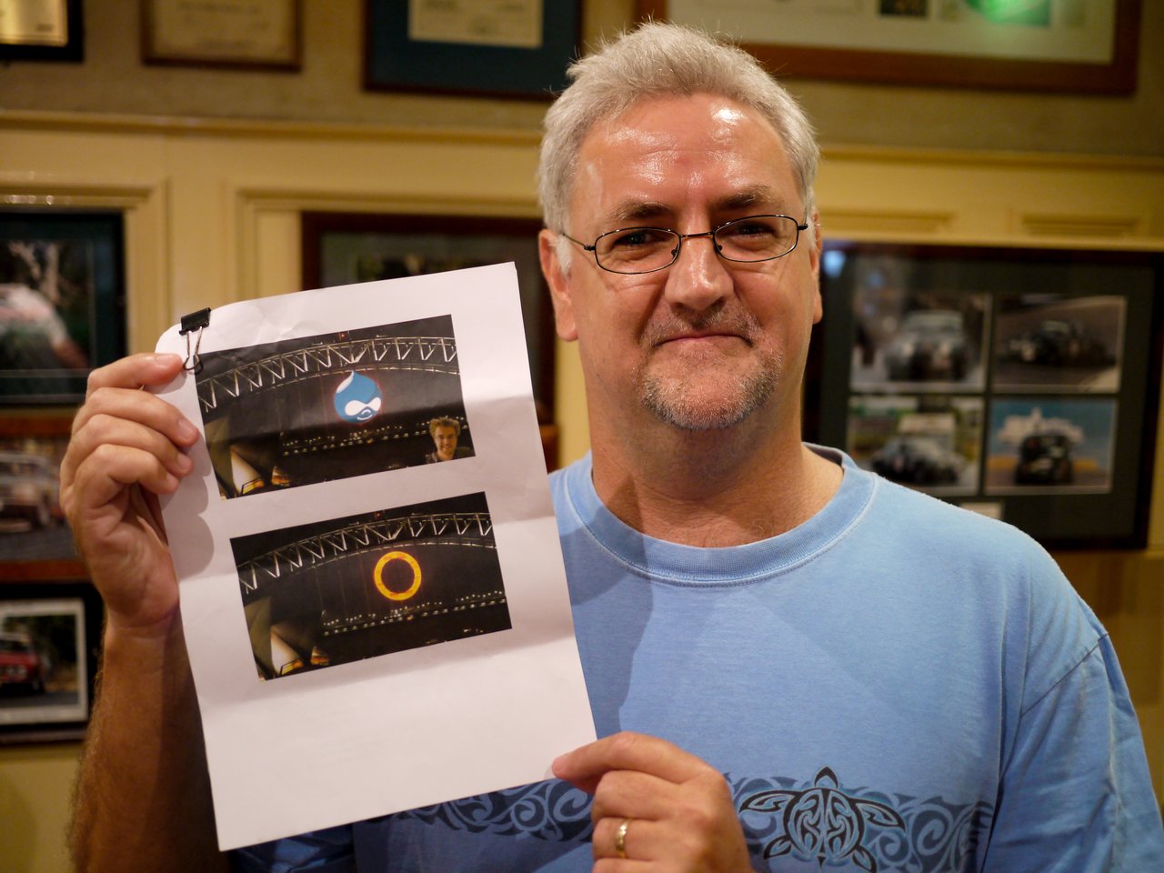 A man in a blue shirt holds a printed sheet showing two edited images of the Sydney Harbour Bridge.