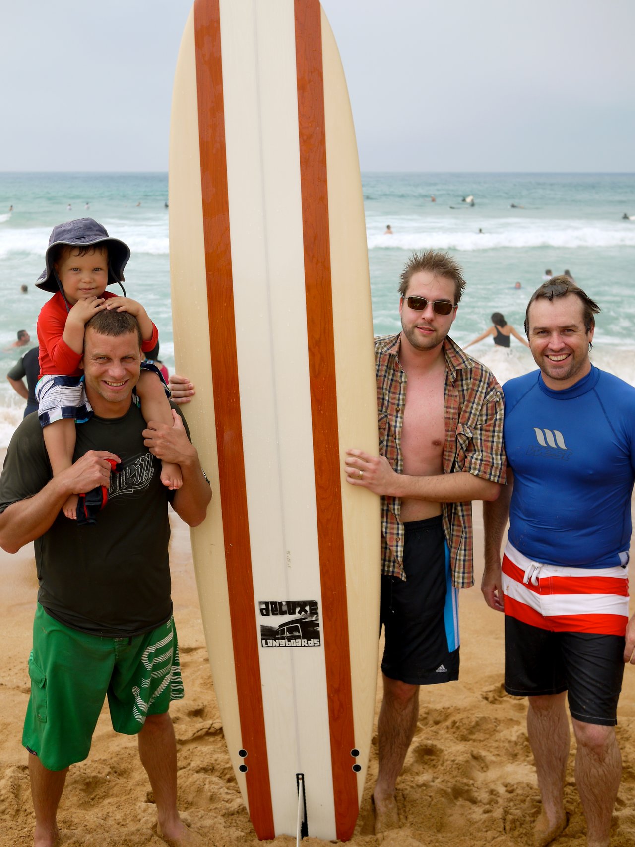 Four men, one carrying a child, stand on a sandy beach holding a long surfboard, with ocean waves behind them.