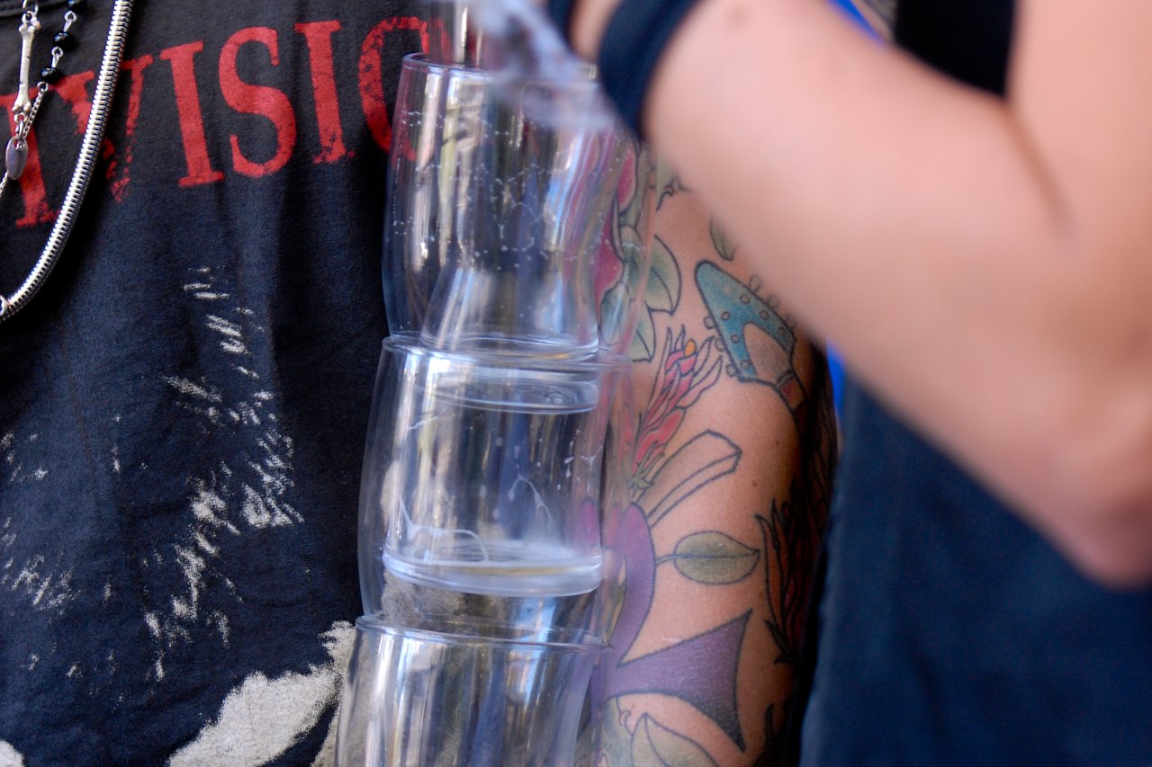 A person balances a stack of clear plastic cups while another person with tattoos moves nearby at a party.