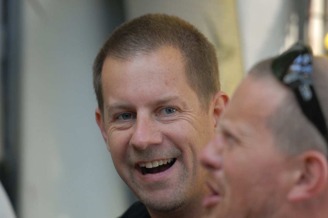 Two men at a party, one smiling and the other talking while wearing sunglasses on his head.