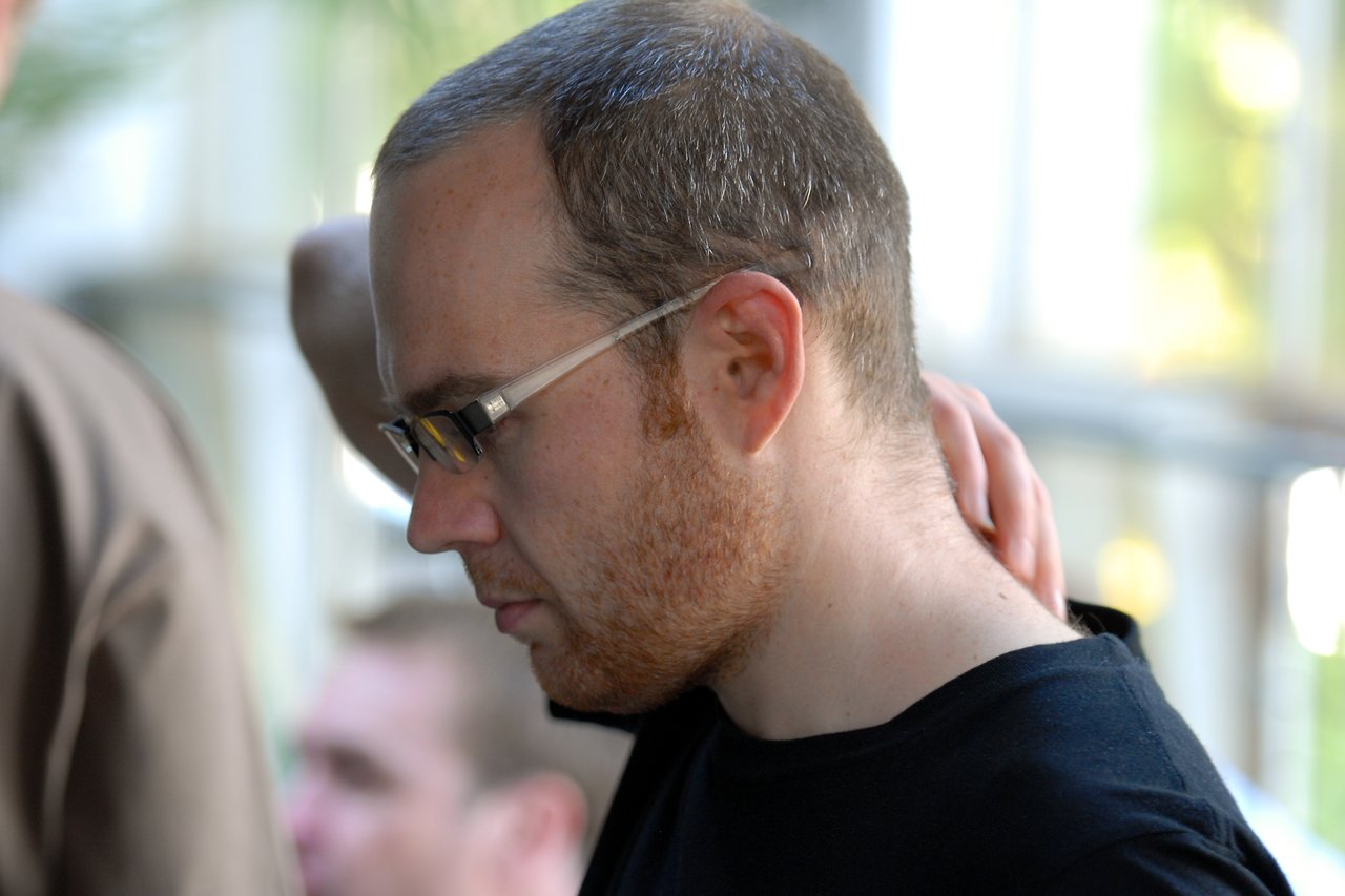 A man wearing glasses and a black shirt touches the back of his neck at a gathering.