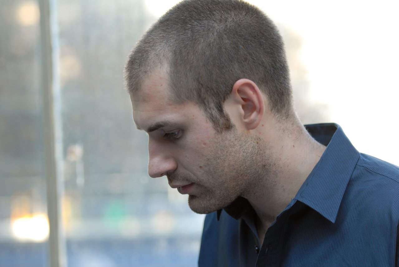 A man in a blue shirt looks down with a serious expression at an indoor or outdoor gathering.