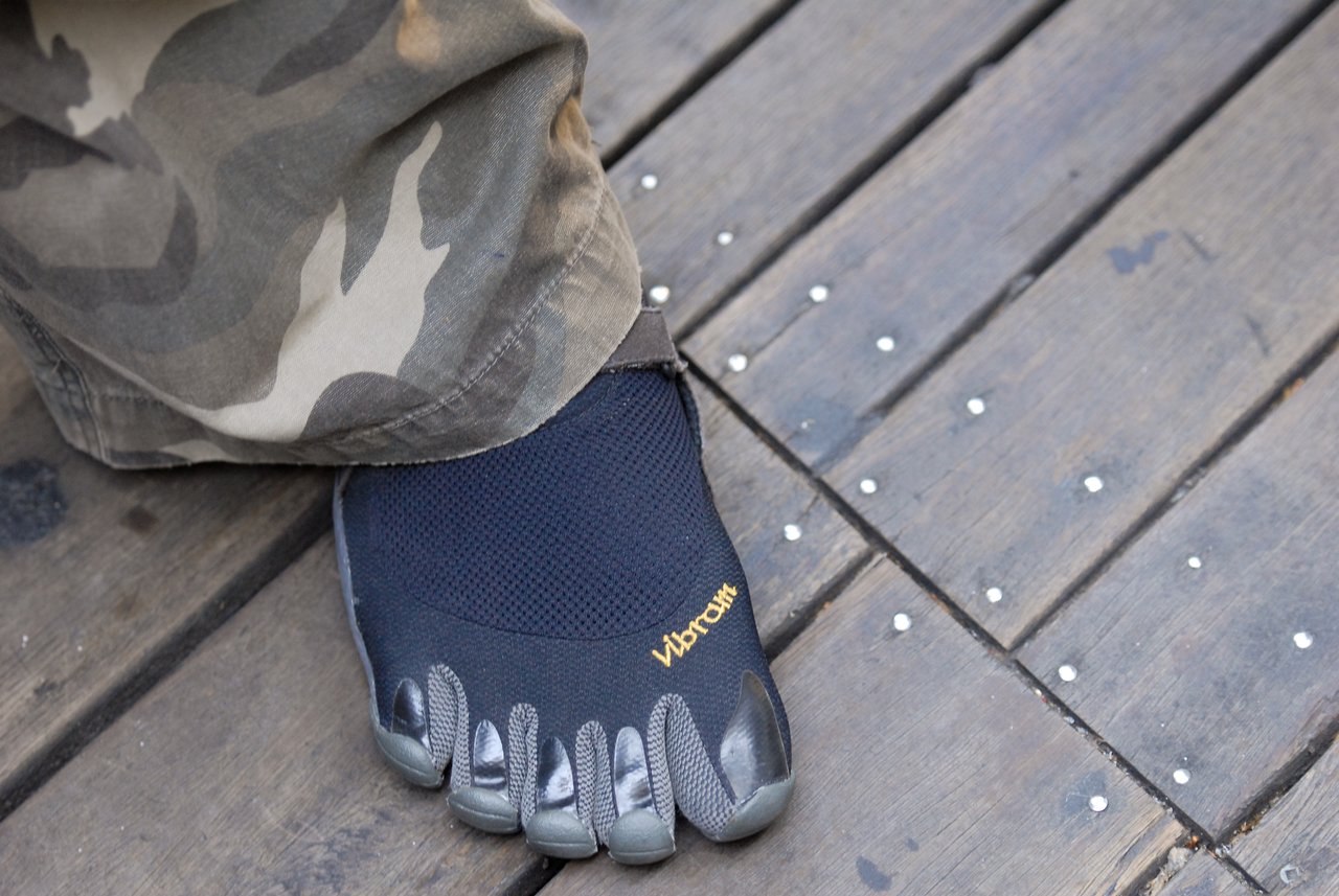 A person wearing camouflage pants and black Vibram five-toe shoes stands on a wooden deck.