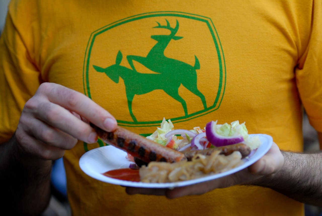 A person in a yellow shirt holds a plate of food and picks up a grilled sausage.
