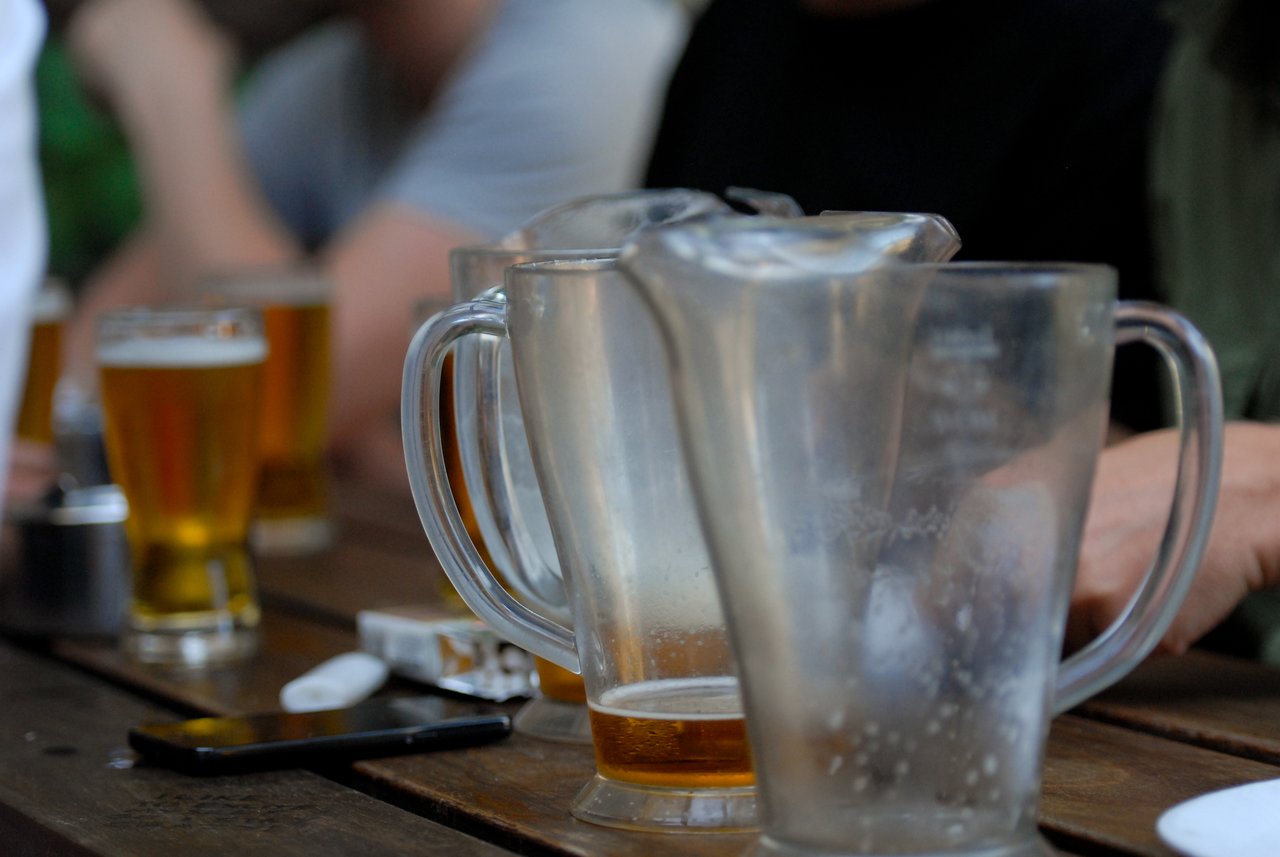Several beer pitchers and glasses on a wooden table, with people sitting and socializing in the background.