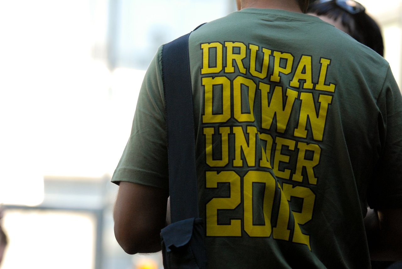 A person wearing a green "Drupal Down Under 2012" T-shirt stands in a crowded party setting.