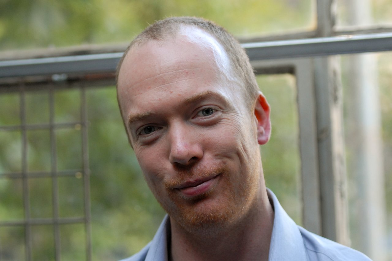 A man with short hair and light facial hair smiles slightly while standing near a window at a party.