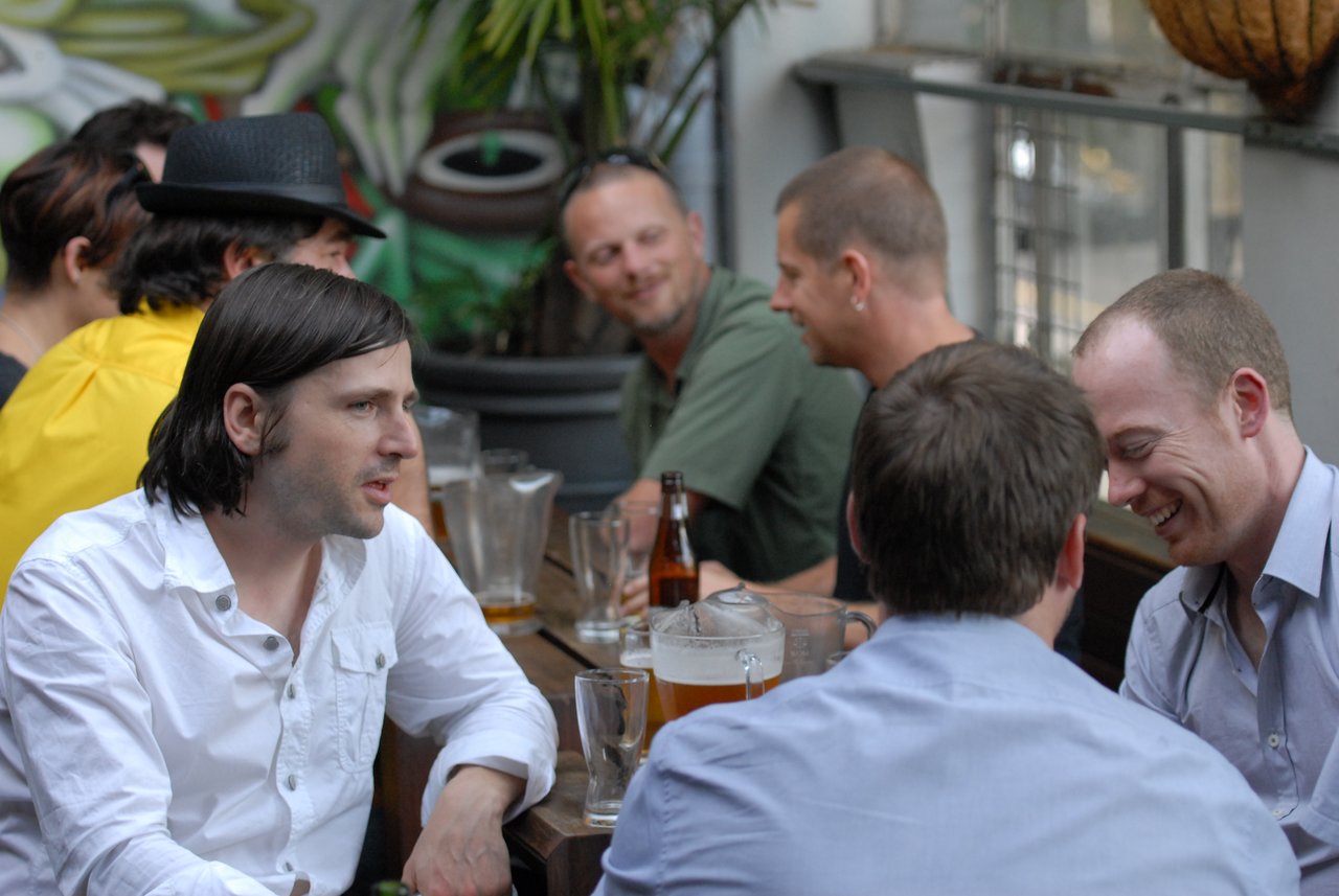 A group of people sit at a table, talking and laughing with drinks in a casual outdoor setting.