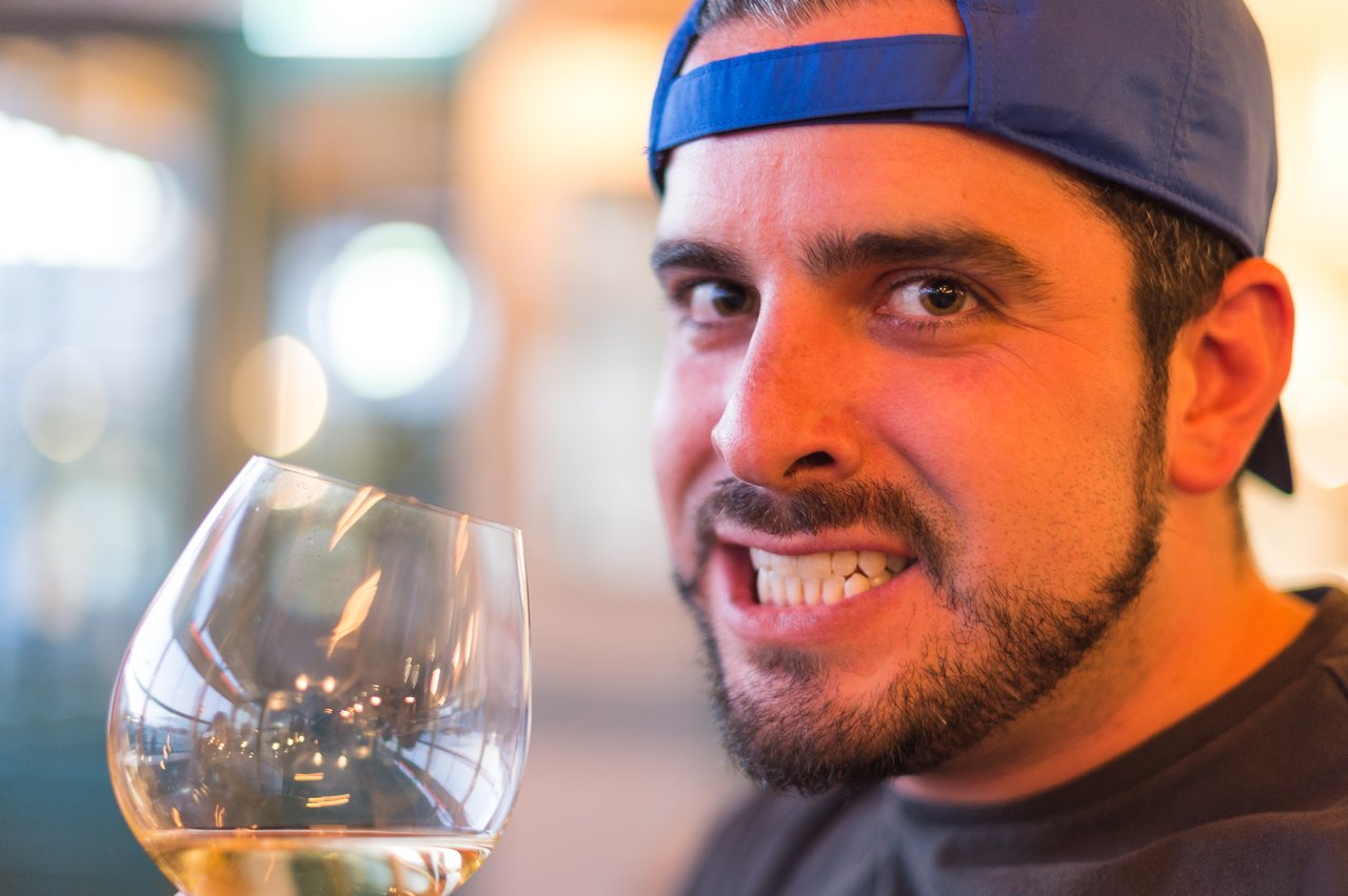 Man wearing a backward blue cap grins while holding a glass of white wine tilted toward the camera.