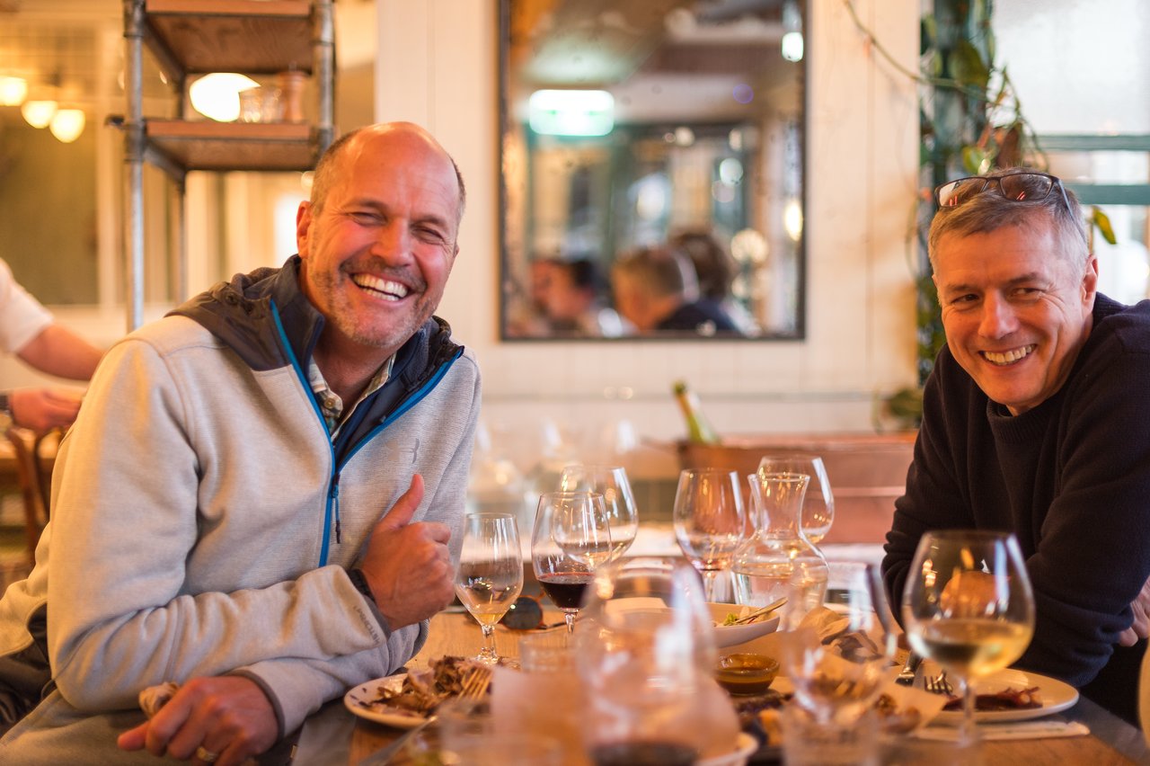 Two men sit at a restaurant table, smiling and enjoying drinks and food.