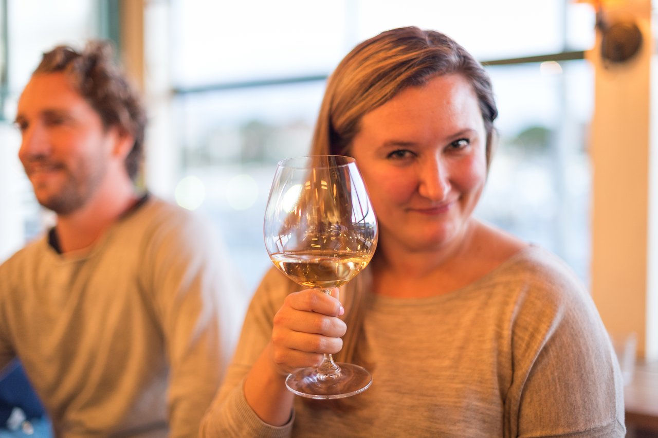 A woman in a gray top holds a glass of white wine and smiles.
