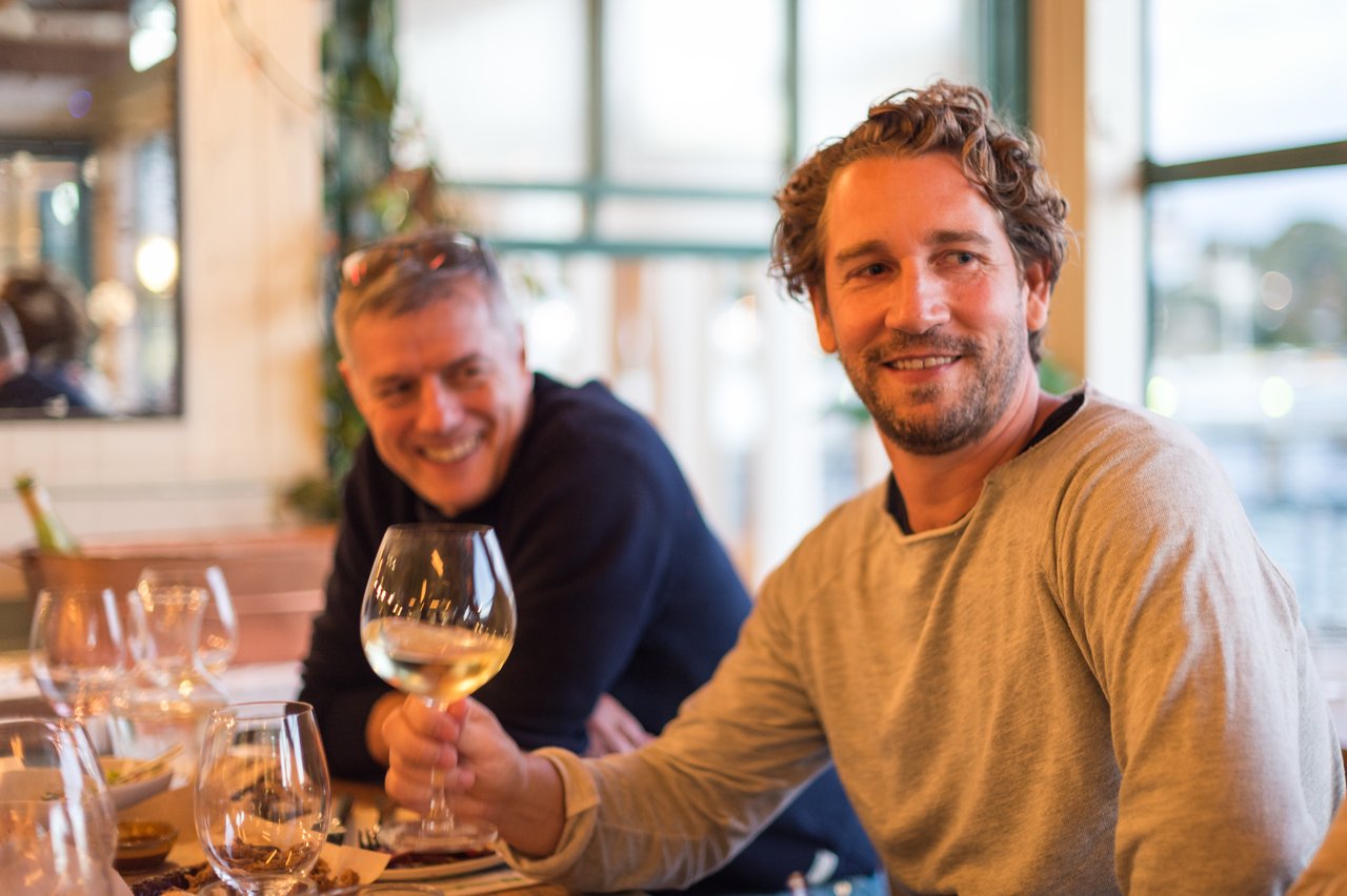 Two men sitting at a restaurant table, one holding a glass of white wine and smiling at the camera.