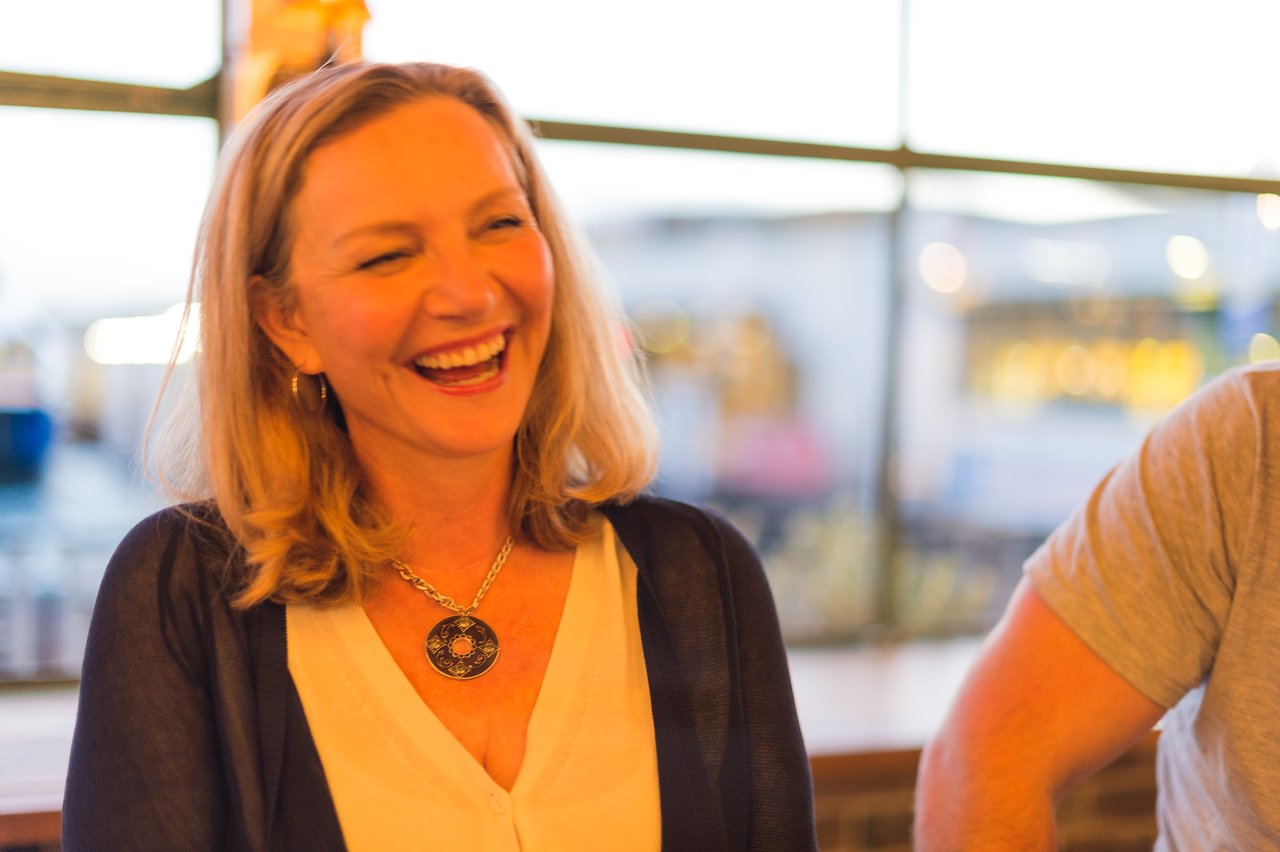 A woman with blonde hair and a necklace laughing in a casual setting.