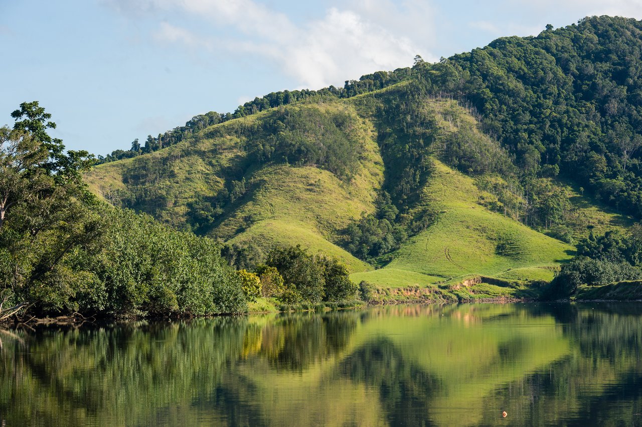 A calm river reflects green hills and trees under a clear sky.