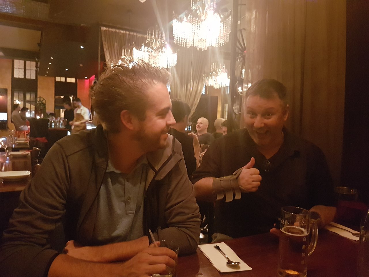 Two men sitting at a restaurant table, one smiling and giving a thumbs-up while the other looks at him.