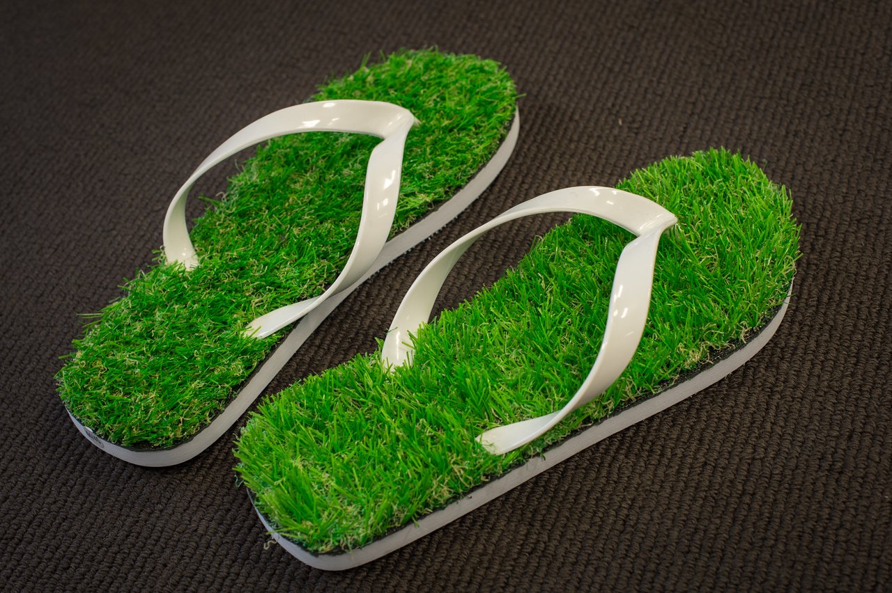 A pair of flip-flops with artificial grass soles and white straps placed on a dark surface.