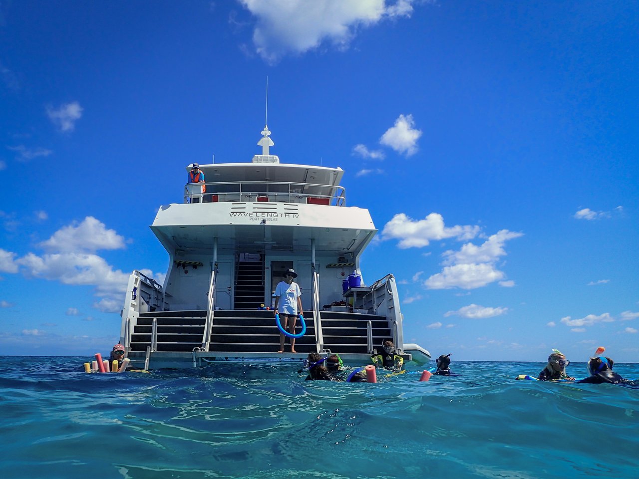 People wearing snorkeling gear float in the water near a boat, while a crew member stands on the steps.