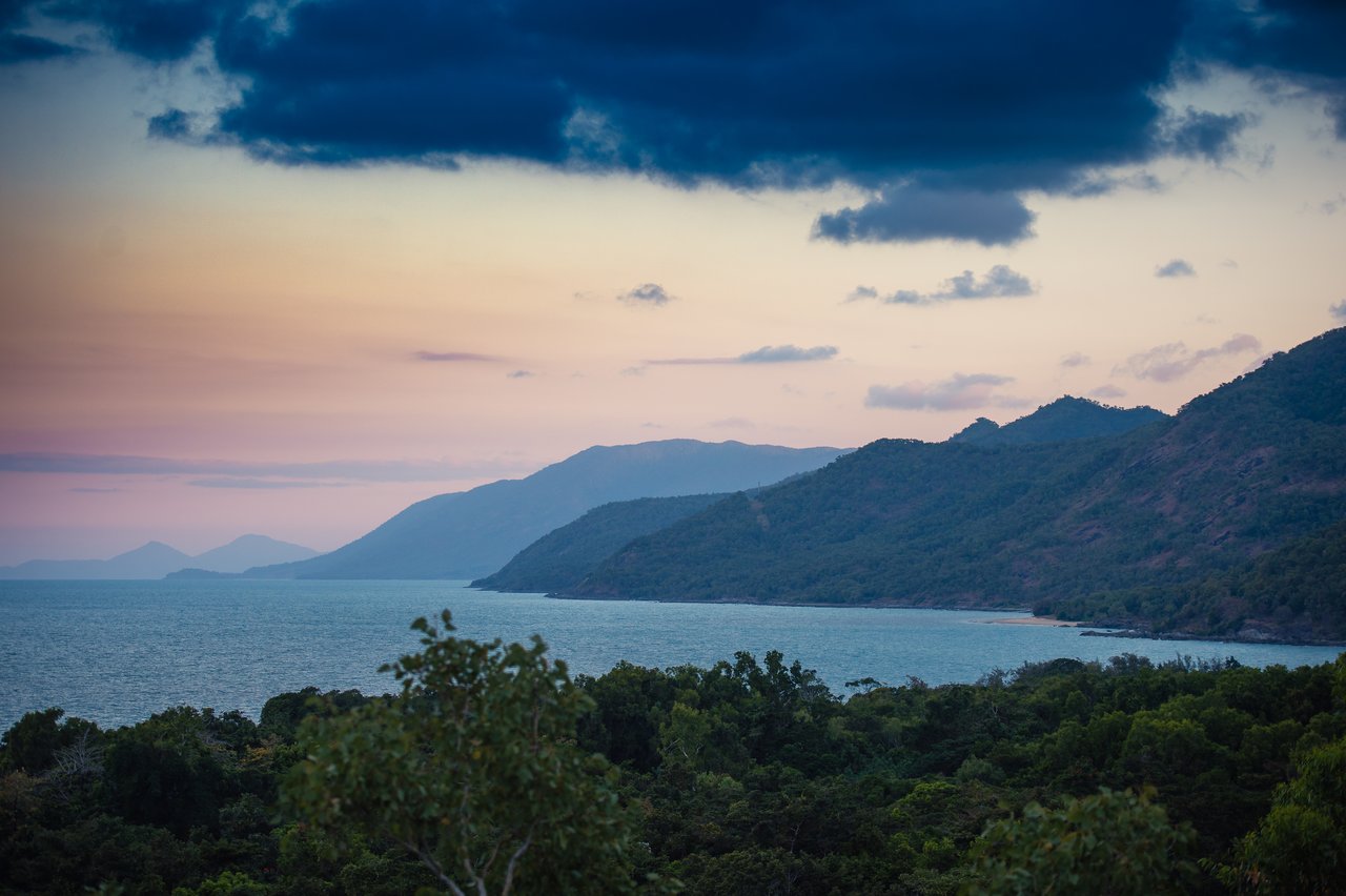A coastal landscape with green mountains, calm blue water, and a colorful sky at dusk.