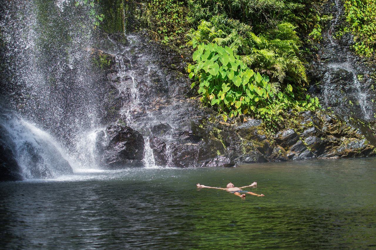 A person floats on their back in a calm pool beneath a waterfall, arms and legs stretched out.