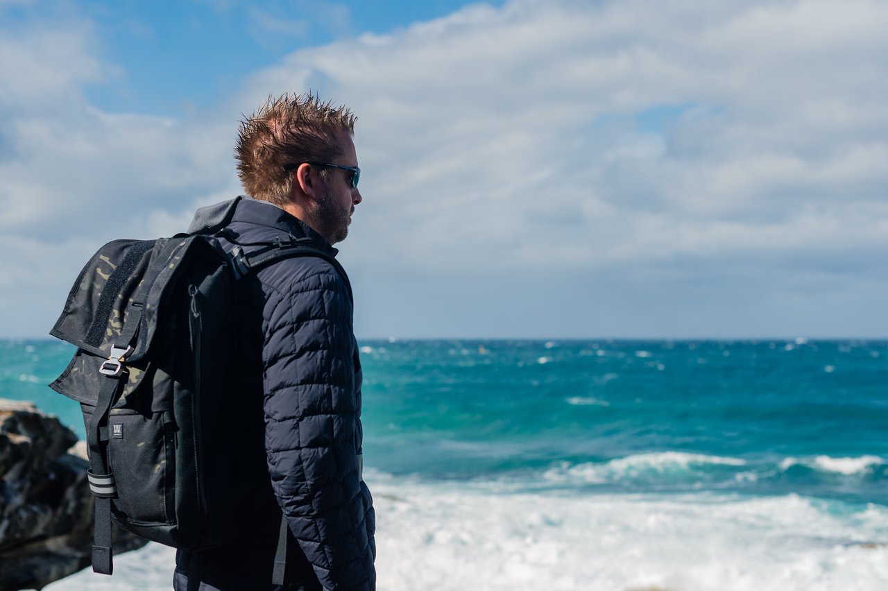 A man wearing a backpack and sunglasses stands near the ocean, looking at the waves.