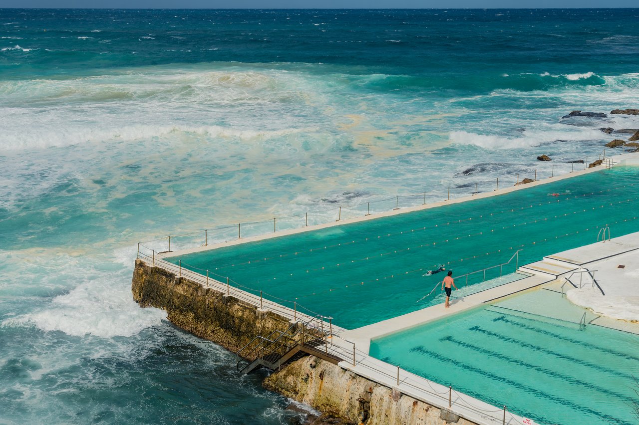 A person walks next to an outdoor swimming pool beside the ocean.