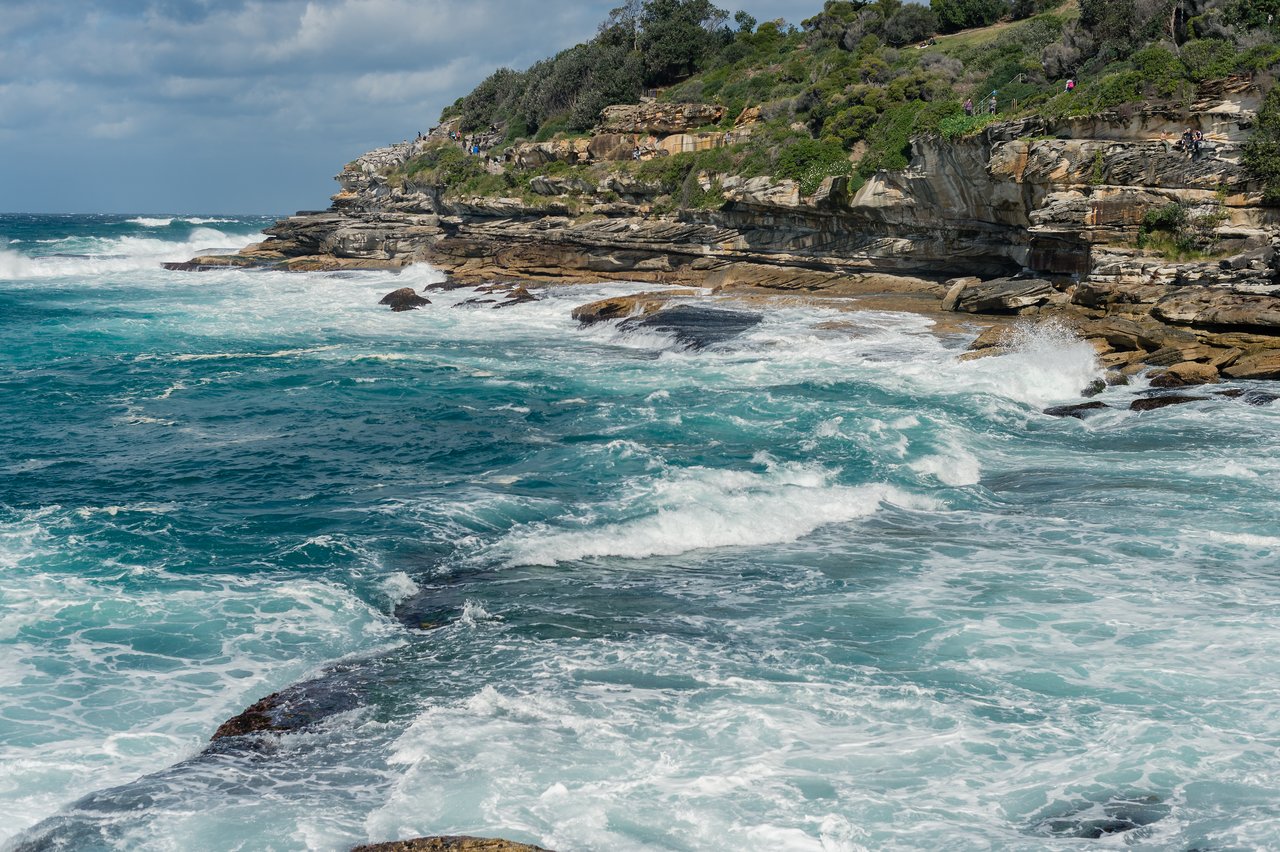 Waves crash against rocky cliffs as people walk along a coastal trail above the shoreline.