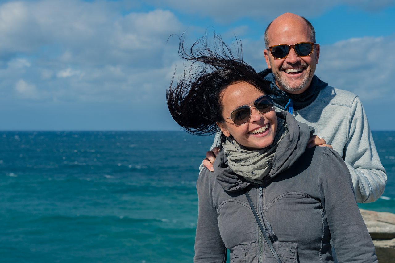 Smiling people wearing sunglasses standing close together with the wind blowing the woman's hair.